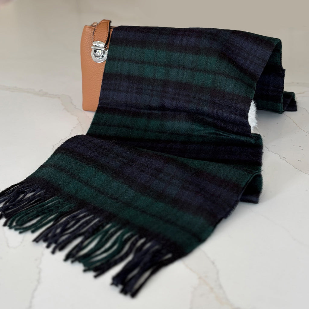 A dark green and blue tartan fringed cashmere scarf draped over a light-colored marble surface, next to a tan leather clutch with a silver clasp.