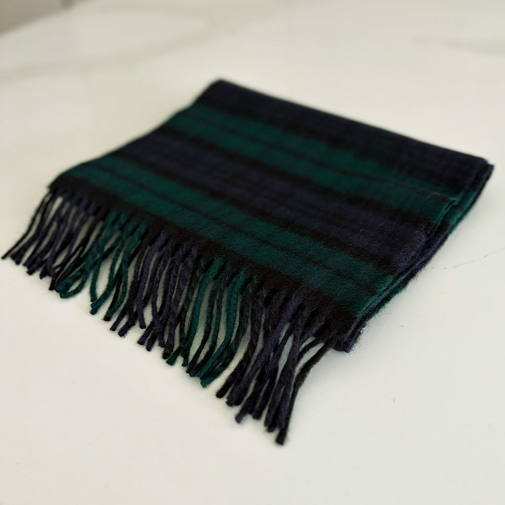 A folded tartan black watch scarf with dark navy and deep green horizontal bands and fringed edges, displayed on a light off-white surface.