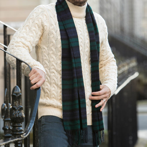 A man wearing a cream cable-knit sweater and a dark green and navy blue tartan cashmere scarf, standing outdoors near a black metal railing.