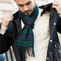 Man wearing a black coat, white sweater, and tartan blue and green cashmere scarf outdoors.