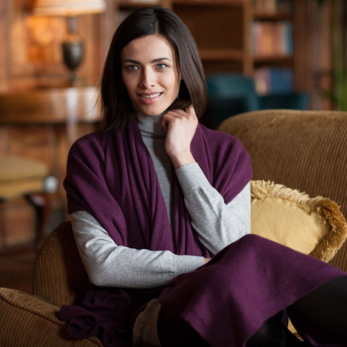 A woman with dark hair sits comfortably in a brown armchair, wearing a deep purple shawl draped over her shoulders over a light gray turtleneck sweater. The background shows a softly blurred warm-toned living room interior with bookshelves and a lamp.