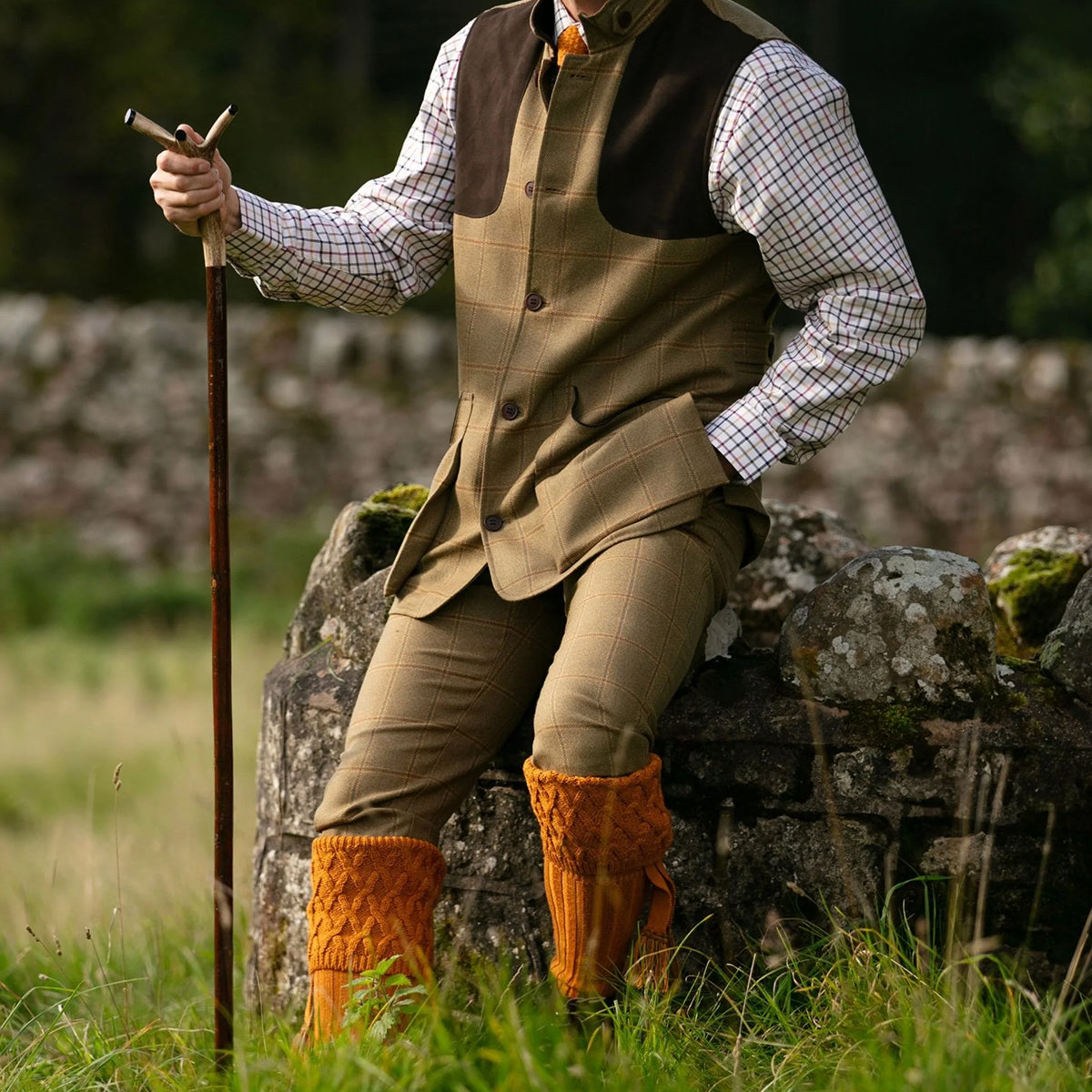A person outdoors wearing a brown and beige plaid waistcoat and matching trousers over a white check shirt, with bright orange cable knit wool socks, seated on a stone wall and holding a walking staff.
