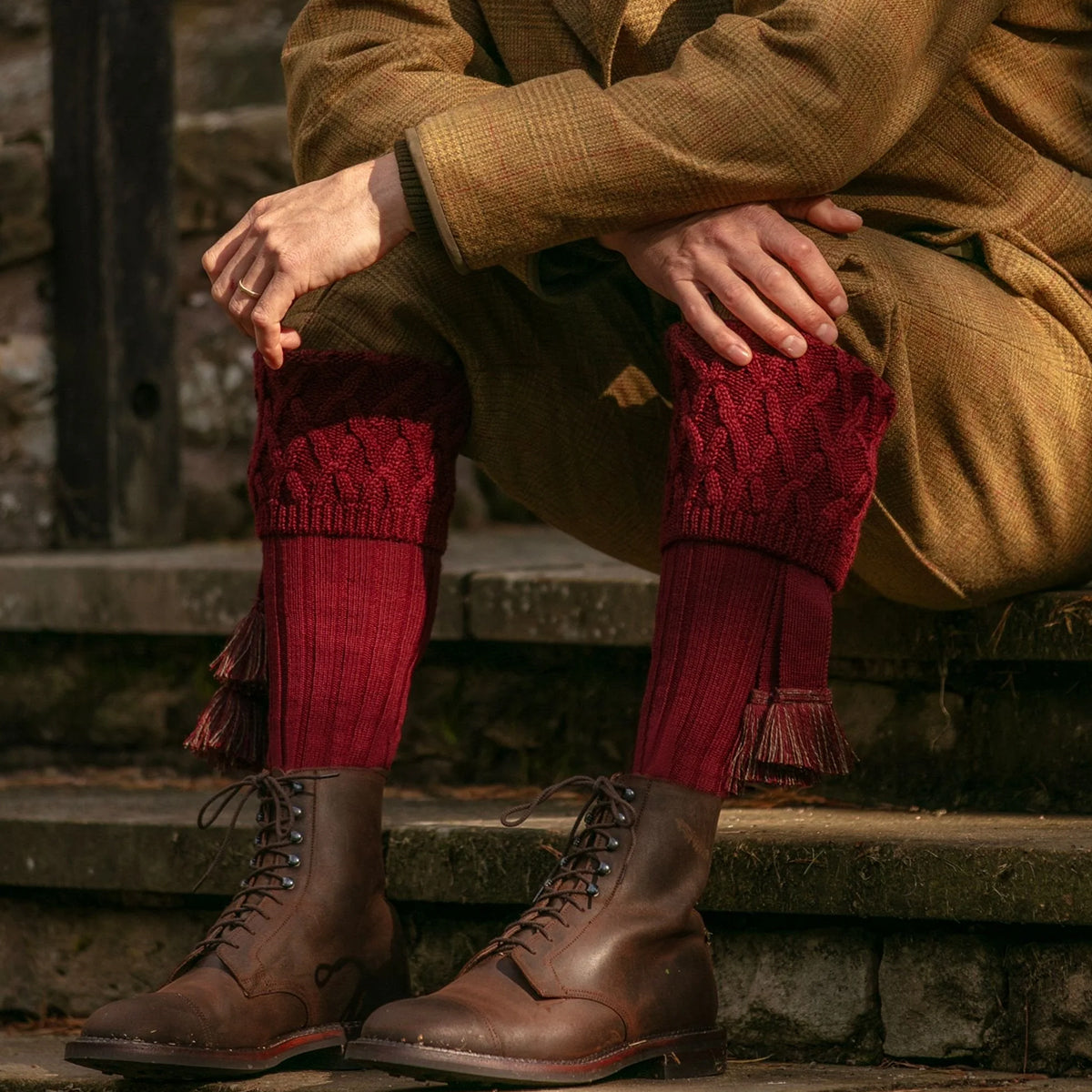 A person seated on stone steps wearing burgundy cable-knit merino wool socks , brown lace-up boots, and a tan coat in an outdoor rustic setting.