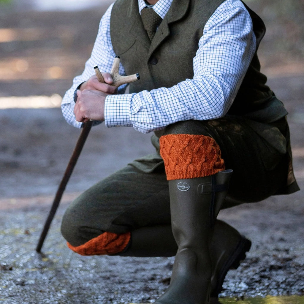 A man in traditional hunting attire crouches outdoors near water, wearing an olive green waistcoat, matching trousers, dark rubber boots, and rust-orange knit wool socks over a light blue gingham shirt.