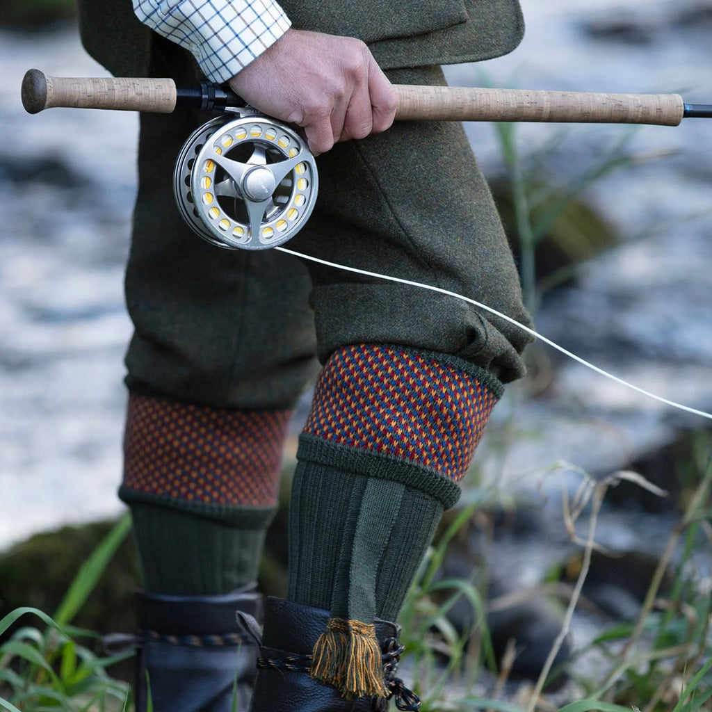 A close-up lifestyle shot of a fly fisherman holding a wooden fly rod with a silver fly reel. The person is wearing traditional fly-fishing attire including olive green tweed trousers with patterned spruce green knee high socks with a colourful birdseye border at the top.