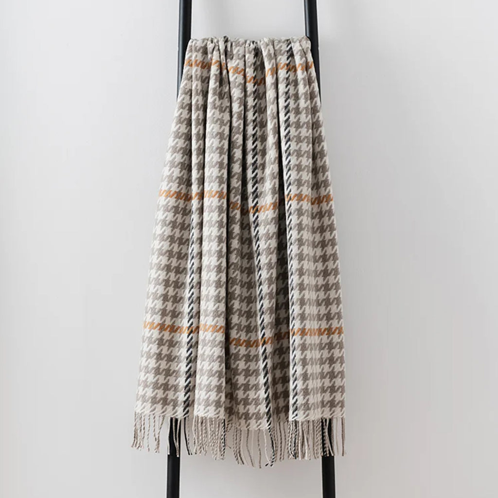 A woven lambswool throw blanket with a houndstooth pattern in beige, grey, and black, featuring thin horizontal rust-orange stripes and fringed edges, draped over a black metal stand against a light grey wall.