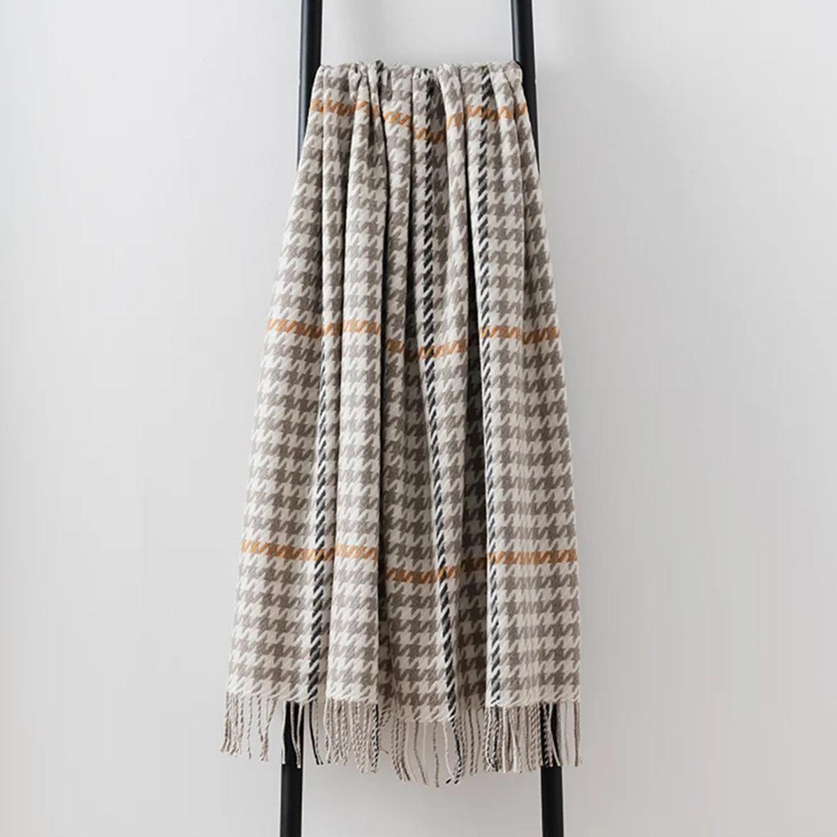A woven lambswool throw blanket with a houndstooth pattern in beige, grey, and black, featuring thin horizontal rust-orange stripes and fringed edges, draped over a black metal stand against a light grey wall.