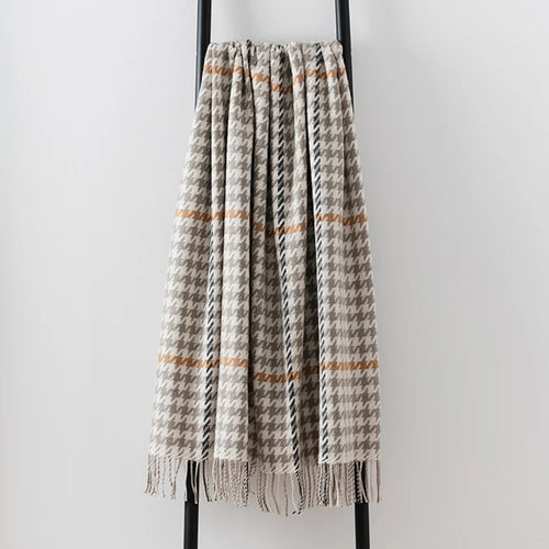 A woven lambswool throw blanket with a houndstooth pattern in beige, grey, and black, featuring thin horizontal rust-orange stripes and fringed edges, draped over a black metal stand against a light grey wall.