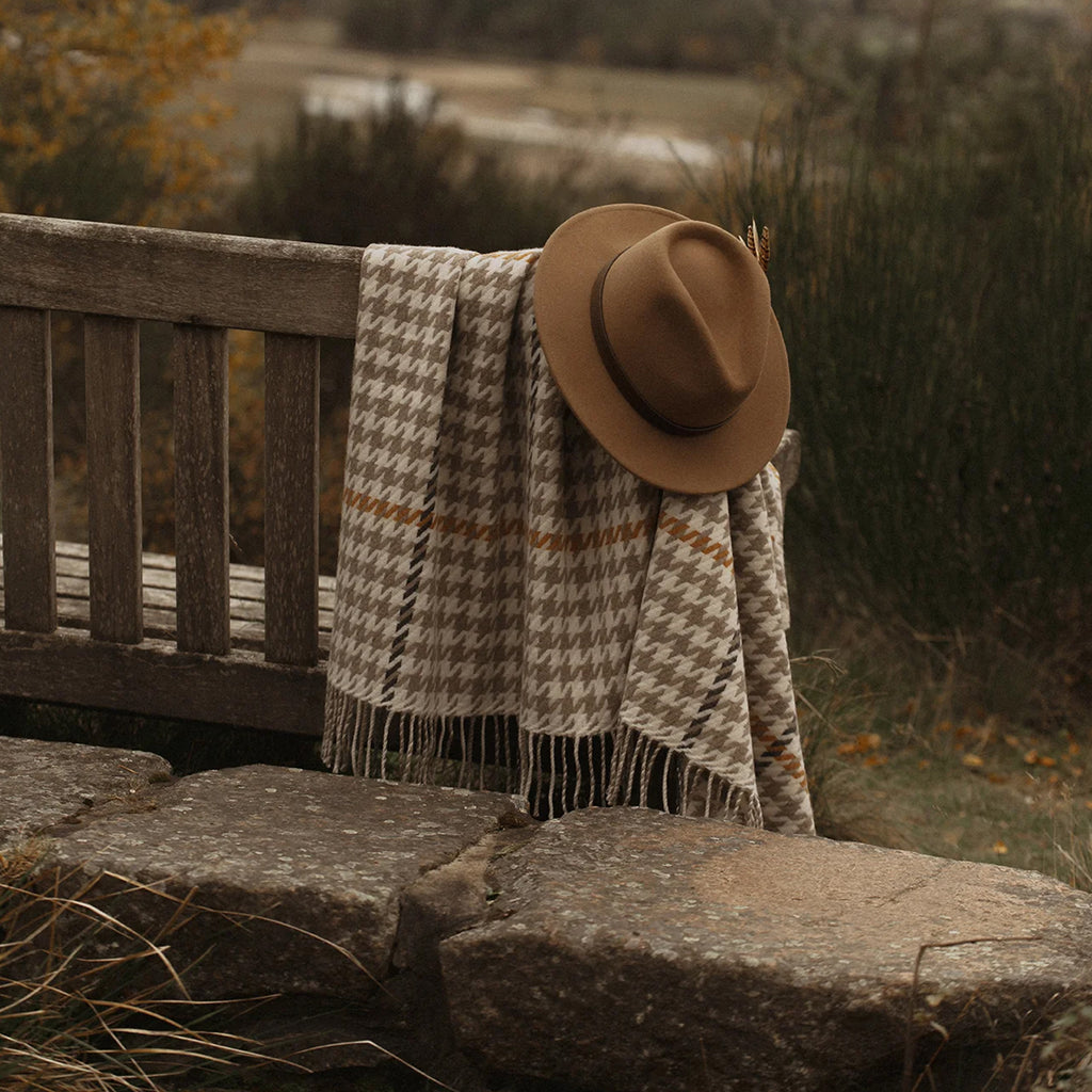 A brown felt hat and a beige and brown houndstooth patterned fringed merino throw blanket are draped over a rustic wooden bench in an outdoor setting with stone steps and a blurred natural background.