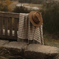 A brown felt hat and a beige and brown houndstooth patterned fringed merino throw blanket are draped over a rustic wooden bench in an outdoor setting with stone steps and a blurred natural background.