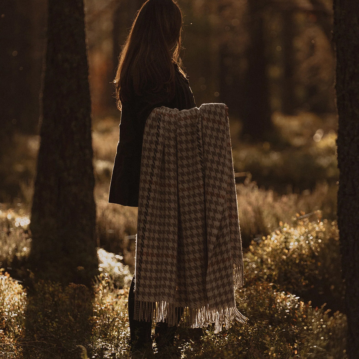 A woman stands in a sunlit forest holding a beige and brown houndstooth patterned fringed throw blanket.