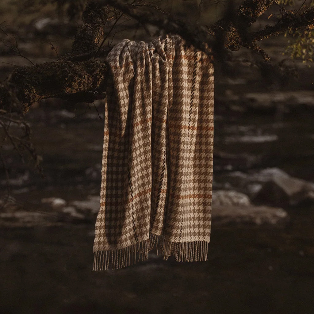 A woven throw with a brown and beige houndstooth pattern and fringe, hanging from a tree branch in a dark, outdoor forest setting.