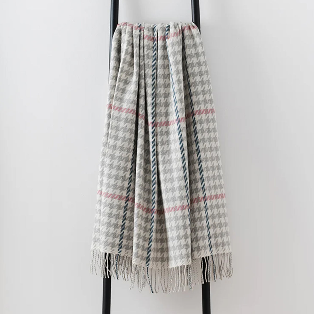 A woven lambswool throw blanket with a gray and white houndstooth pattern, accented by pink overcheck, draped over a black metal stand against a light gray background.