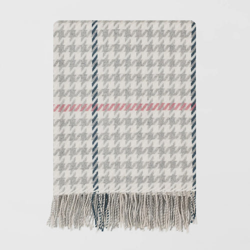 A woven merino throw blanket with a white and silver gray houndstooth pattern, featuring a horizontal pink stripe and fringed edges, shown against a light gray background.