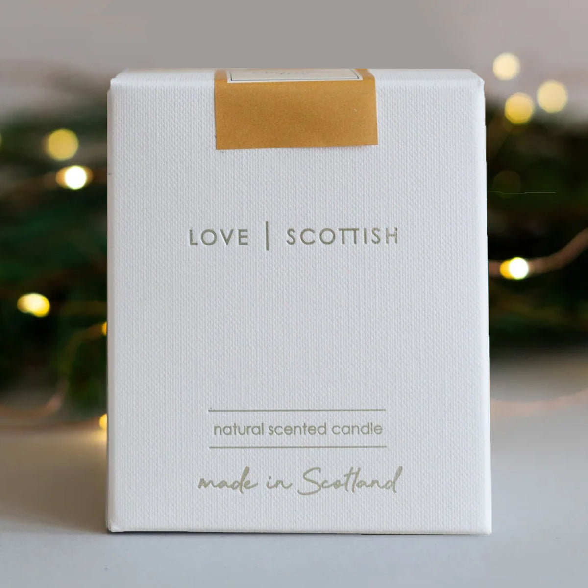 A white square product box for a 'Christmas Tree' scented candle by 'Love Scottish'.  natural scented candle, featuring gold foil text and a gold seal at the top, set against a softly blurred background with warm bokeh lights.