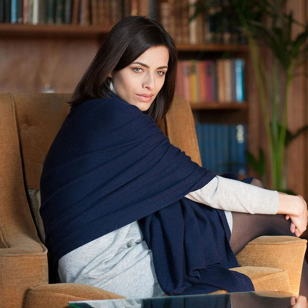 A woman sits in a brown armchair in a cozy indoor setting, wrapped in a dark navy blue shawl or wrap over a light gray top. Bookshelves and a plant are visible in the background.