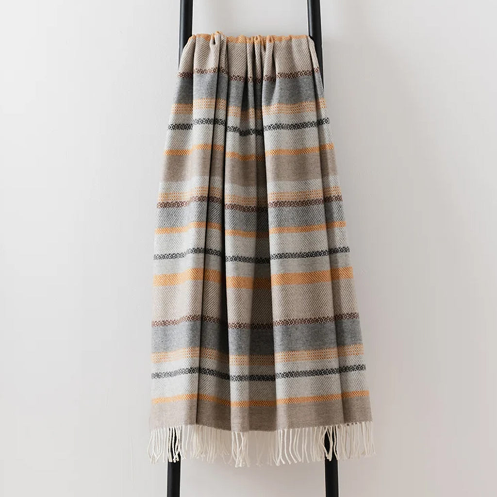 A striped woven fair Isle throw blanket with fringe edge, hanging from a black metal stand against a plain off-white wall. The blanket features horizontal stripes in beige, grey, brown, and orange.