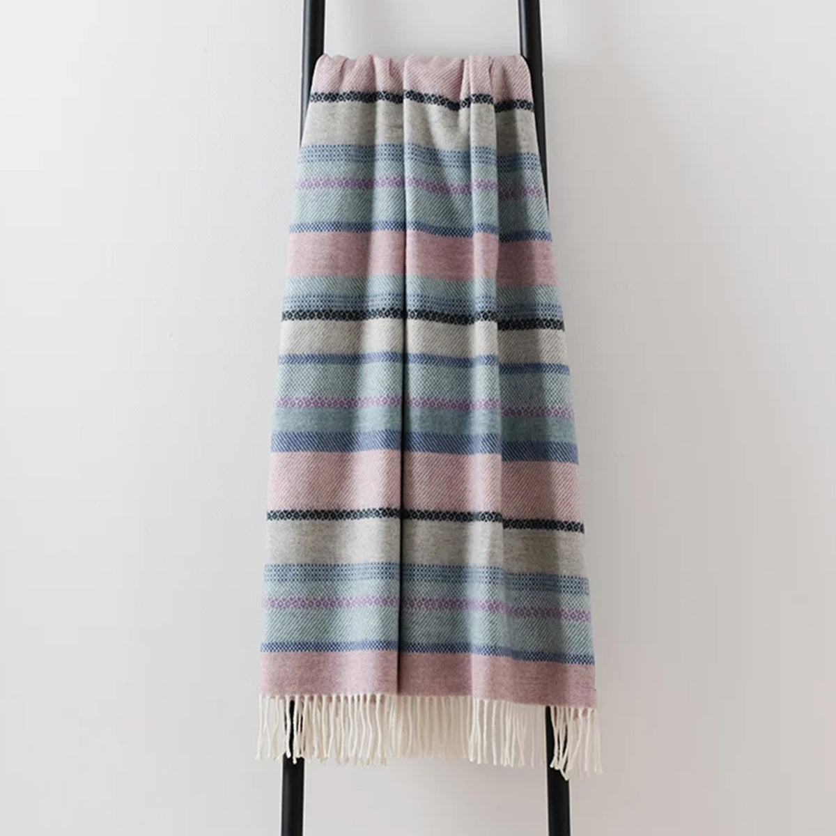 A lambswool merino throw with a fair isle design in muted blue, pink and grey, draped over a black ladder with a pale off-white wall behind.