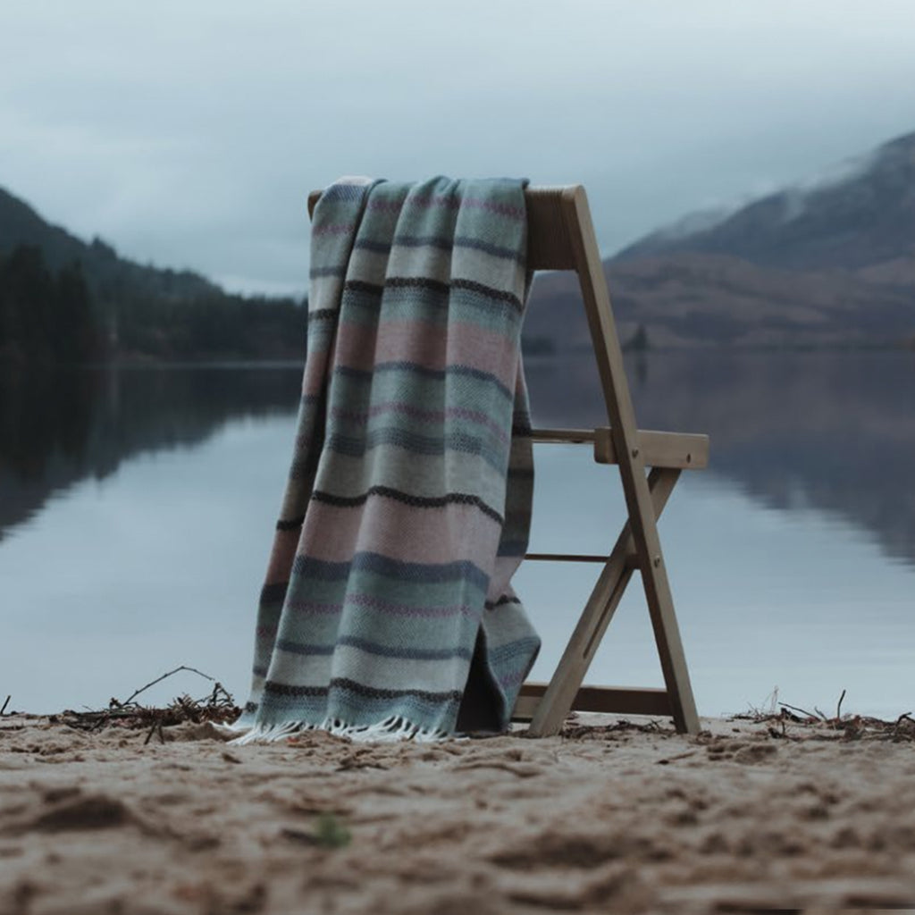 A luxurious Fair Isle Remony lambswool throw draped over a wooden deck chair on a quiet Scottish beach. The hills behind create a tranquil backdrop.