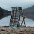 A luxurious Fair Isle Remony lambswool throw draped over a wooden deck chair on a quiet Scottish beach. The hills behind create a tranquil backdrop.
