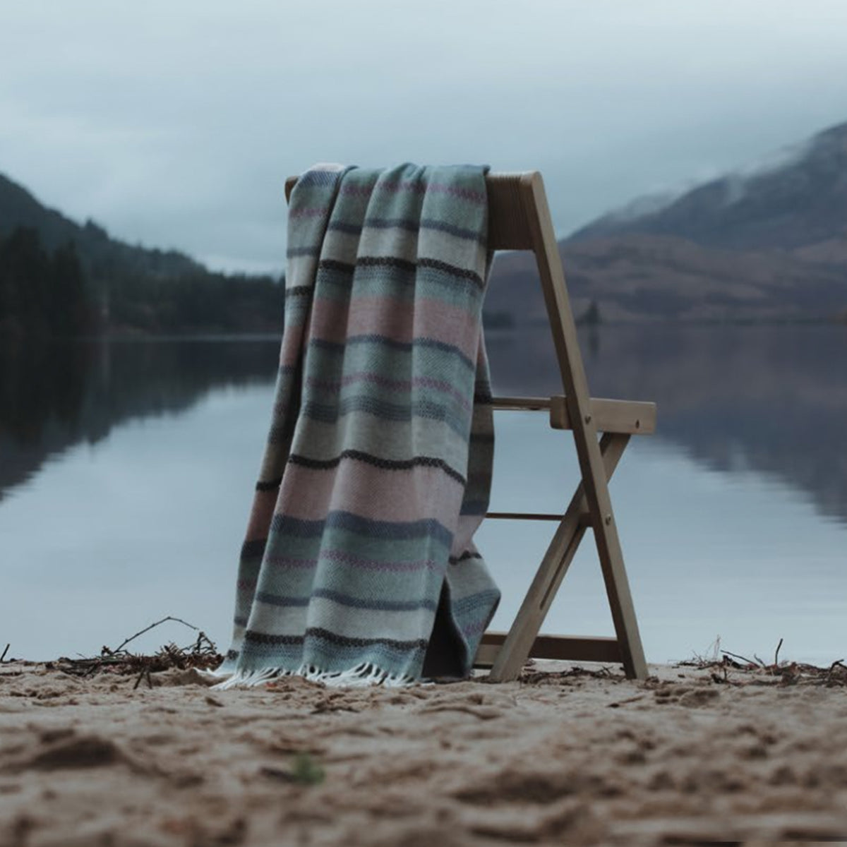 A luxurious Fair Isle Remony lambswool throw draped over a wooden deck chair on a quiet Scottish beach. The hills behind create a tranquil backdrop.