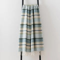 A striped woven lambswool throw blanket with fringe, hanging on a black metal stand against a plain white wall. The blanket features horizontal stripes in light blue, navy, cream and pale green.