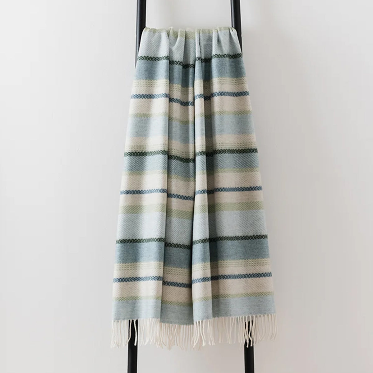 A striped woven lambswool throw blanket with fringe, hanging on a black metal stand against a plain white wall. The blanket features horizontal stripes in light blue, navy, cream and pale green.