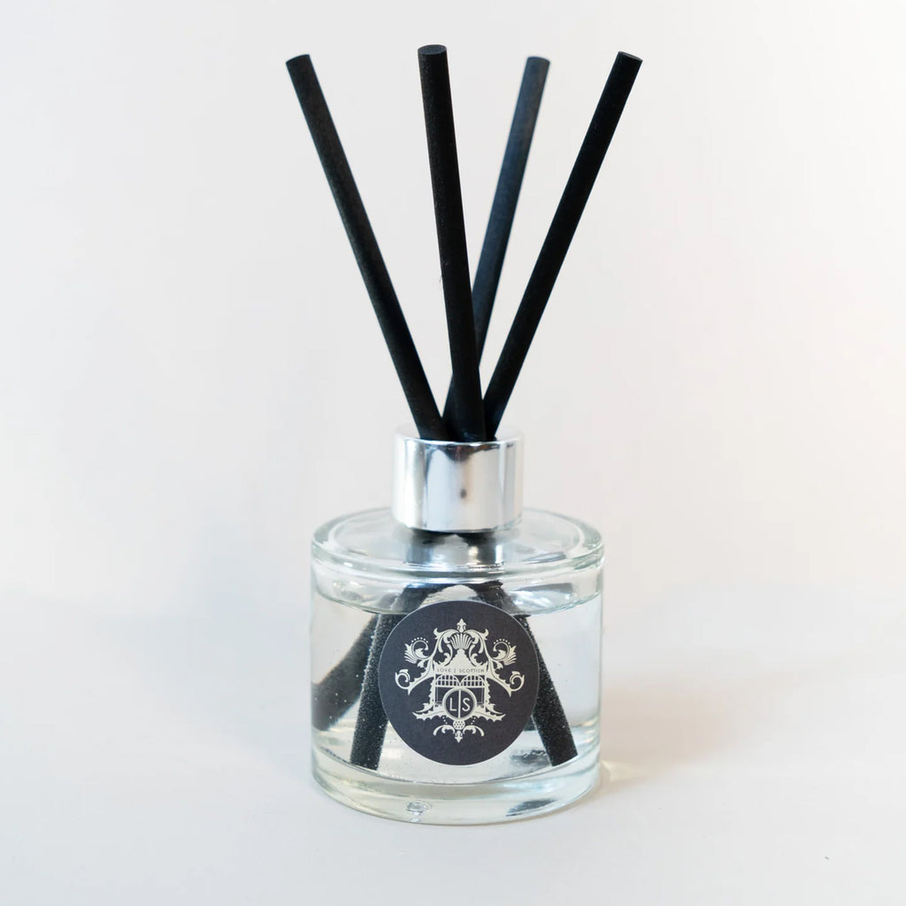 A clear glass reed diffuser bottle with a silver collar and four black reeds, featuring a black circular label with a white crest design, set against a plain white background.
