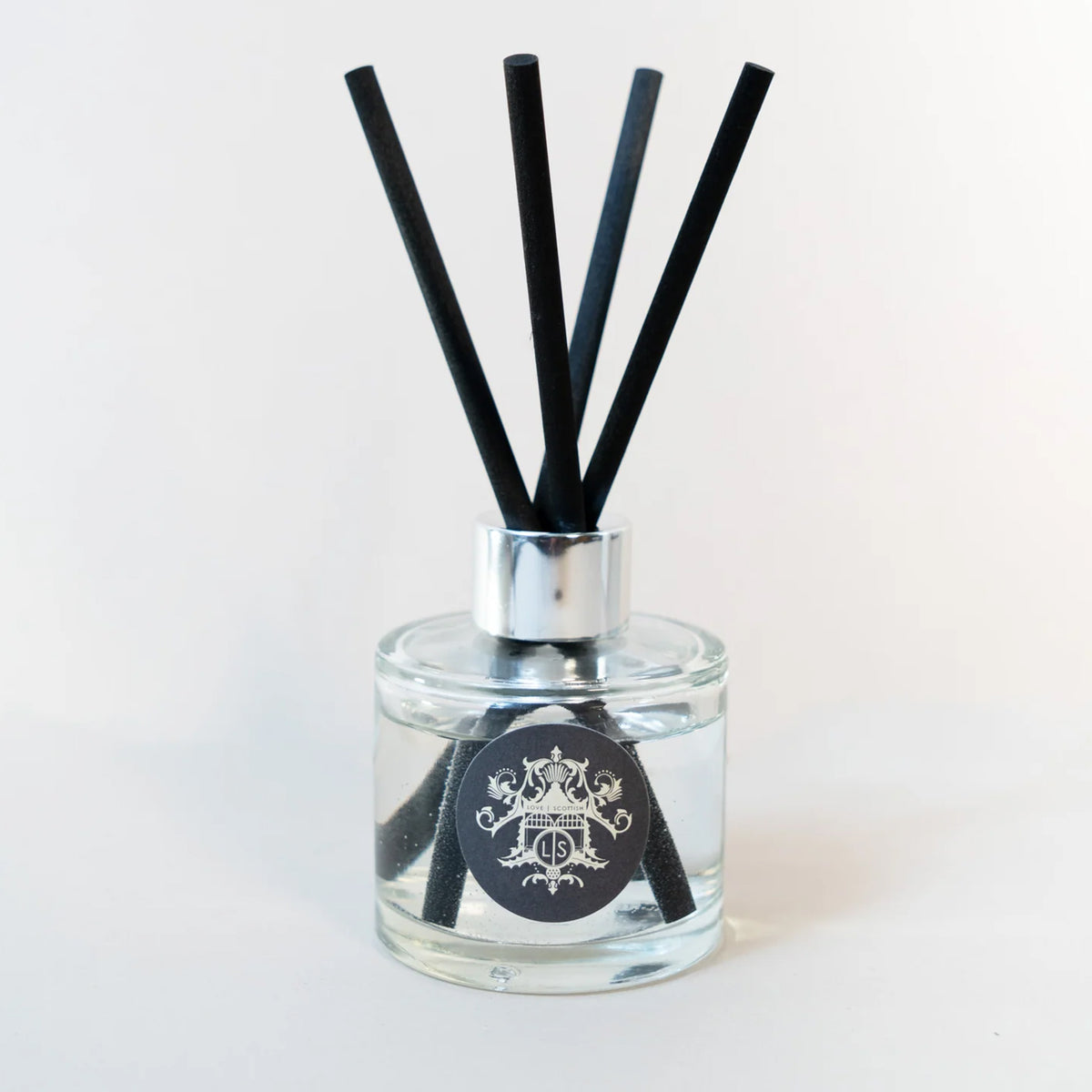 A clear glass reed diffuser bottle with a silver collar and four black reeds, featuring a black circular label with a white crest design, set against a plain white background.