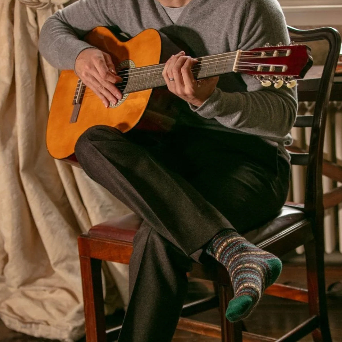 A person sitting on a wooden chair playing a classical acoustic guitar, wearing a fairisle merino woll knit socks with a green toe cap.