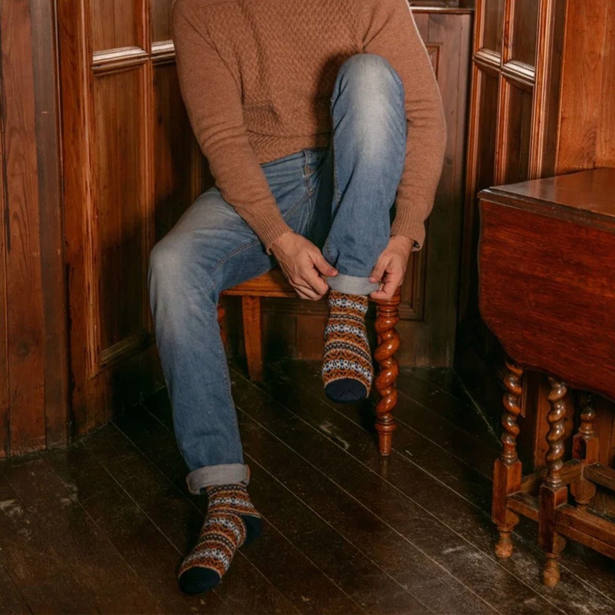 A person sitting on a wooden chair in a rustic room, putting on a fairisle merino wool knitted socks. The person is wearing a brown knit sweater and blue jeans.
