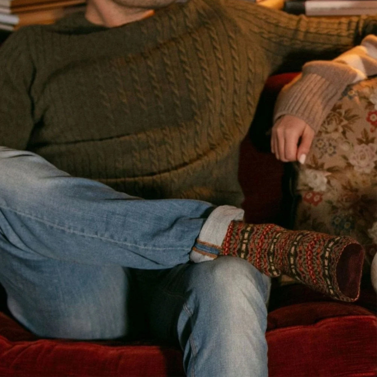A person sitting on a red velvet couch wearing a pair of multicolor fairisle merino wool knit socks in rust brown.