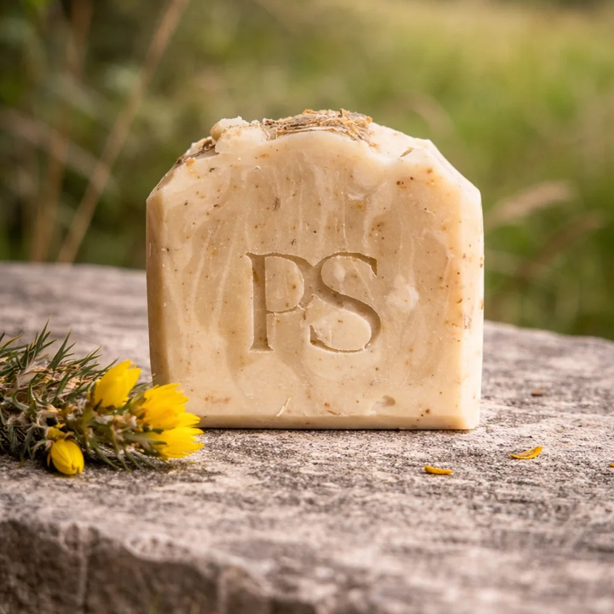 A rectangular bar of handmade soap with a rough, rustic finish and speckled natural inclusions, embossed with the initials 'Purple Sage'. It is displayed on a weathered stone surface next to small yellow flowers, with a soft-focus outdoor green background.