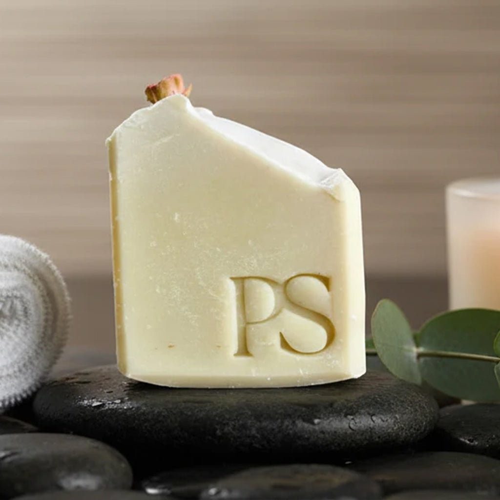 A rectangular bar of pale cream soap with a carved 'Purple Sage' monogram, resting on dark spa stones with a rolled towel and eucalyptus leaves in a spa-like setting.