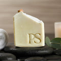 A rectangular bar of pale cream soap with a carved 'Purple Sage' monogram, resting on dark spa stones with a rolled towel and eucalyptus leaves in a spa-like setting.