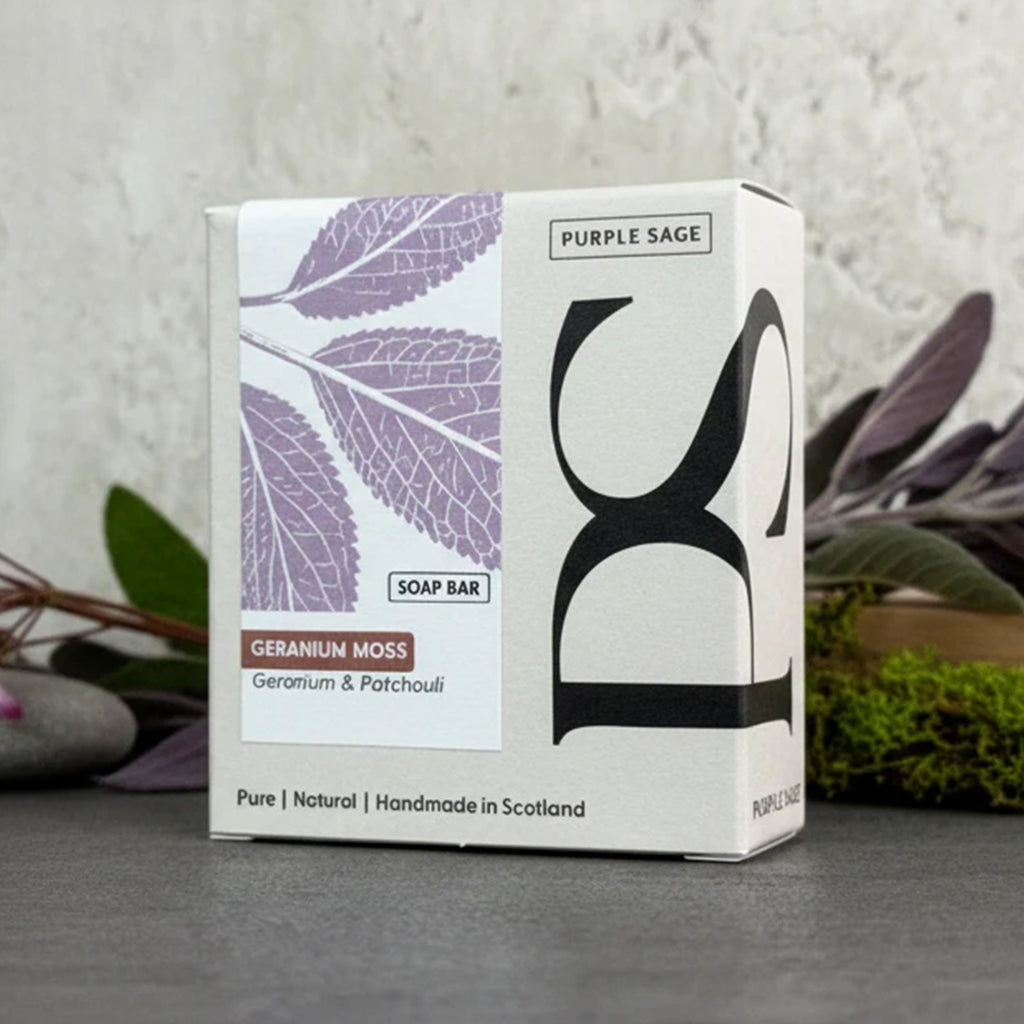 A boxed soap bar product labeled 'Purple Sage' and 'Geranium Moss' sits on a gray surface against a textured light wall, with blurred greenery in the background. The box is white with a lavender leaf print and large black lettering.