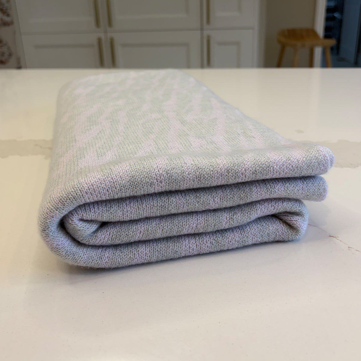 A folded knit blanket with a light grey and pale lilac wavy pattern, resting on a white countertop in a kitchen setting.