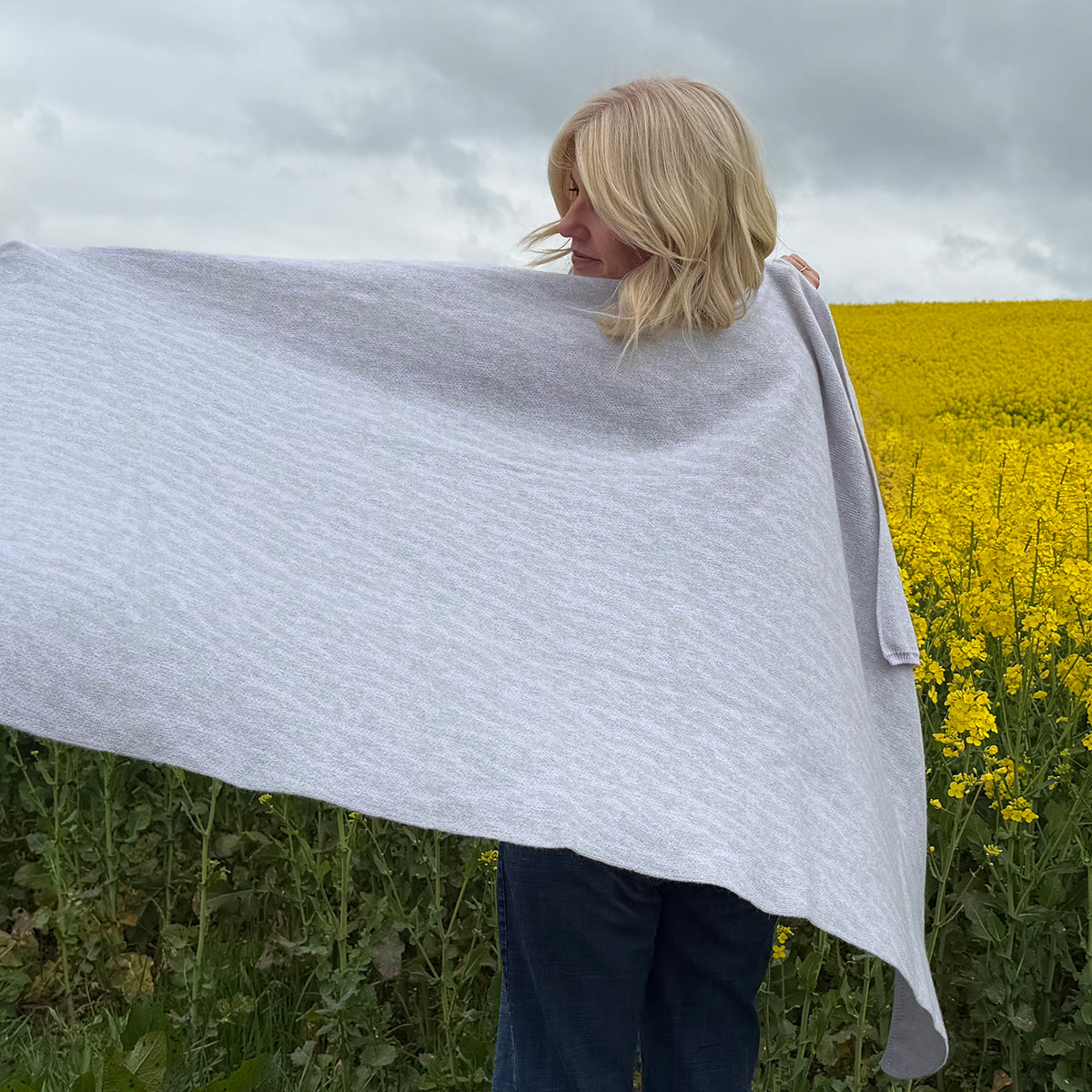 A woman with blonde hair stands in a field of yellow flowers, holding up a large light gray and lilac cashmere shawl. The sky is overcast, and the field is visible in the background.