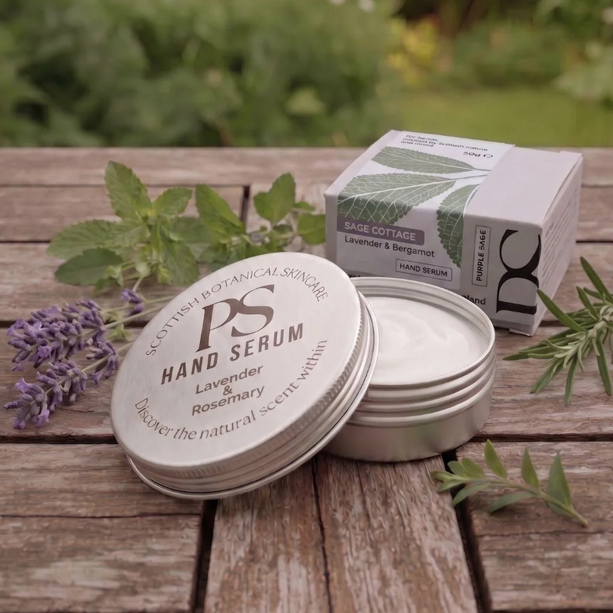 An open round metal tin of Purple Sage Hand Serum and its matching box sit on a rustic wooden table, surrounded by lavender flowers and green herbs. The background is a soft-focus outdoor garden setting.