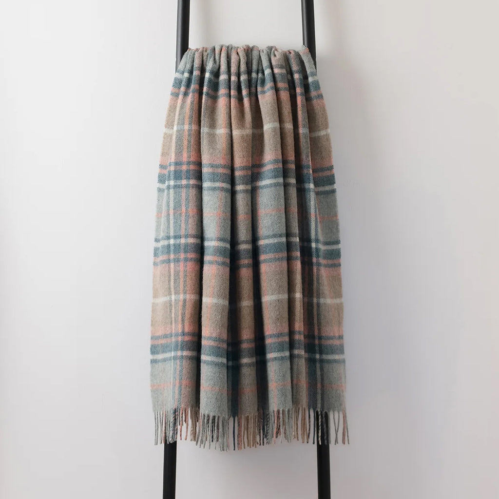 A recycled wool throw draped over a black metal stand - in a pale blue, beige and coral pink shade with an elegant fringe edge.