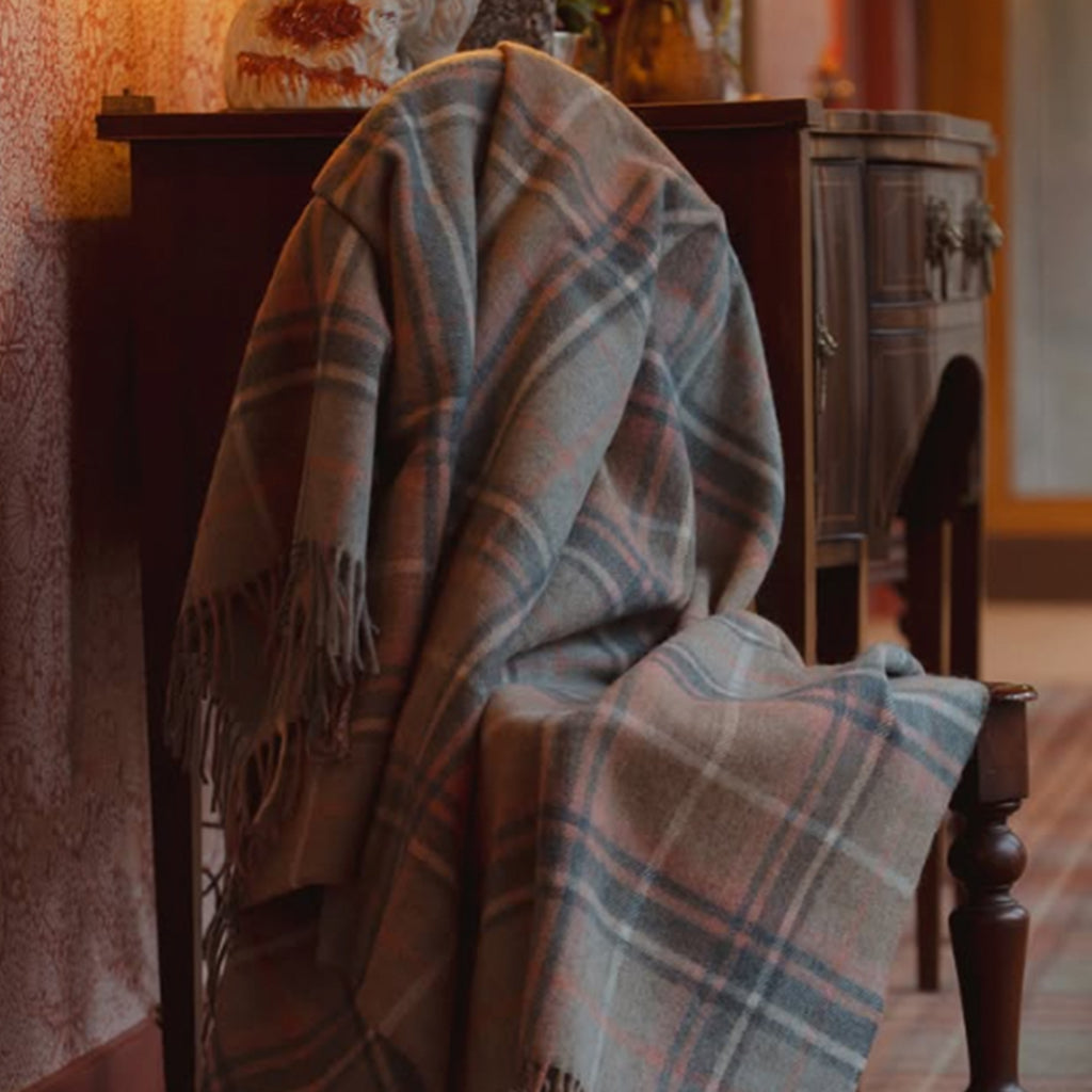 A plaid recycled wool throw blanket draped over a dark wooden chair in a warm, vintage-style room. The blanket features a muted pattern of blue, beige, and dusty pink with fringed edges.