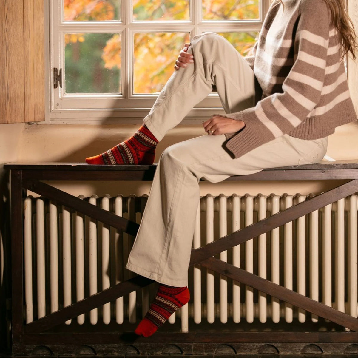 A person sitting on a dark wooden radiator in a cozy room, wearing a bright orange merino wool fairisle patterned knit socks.