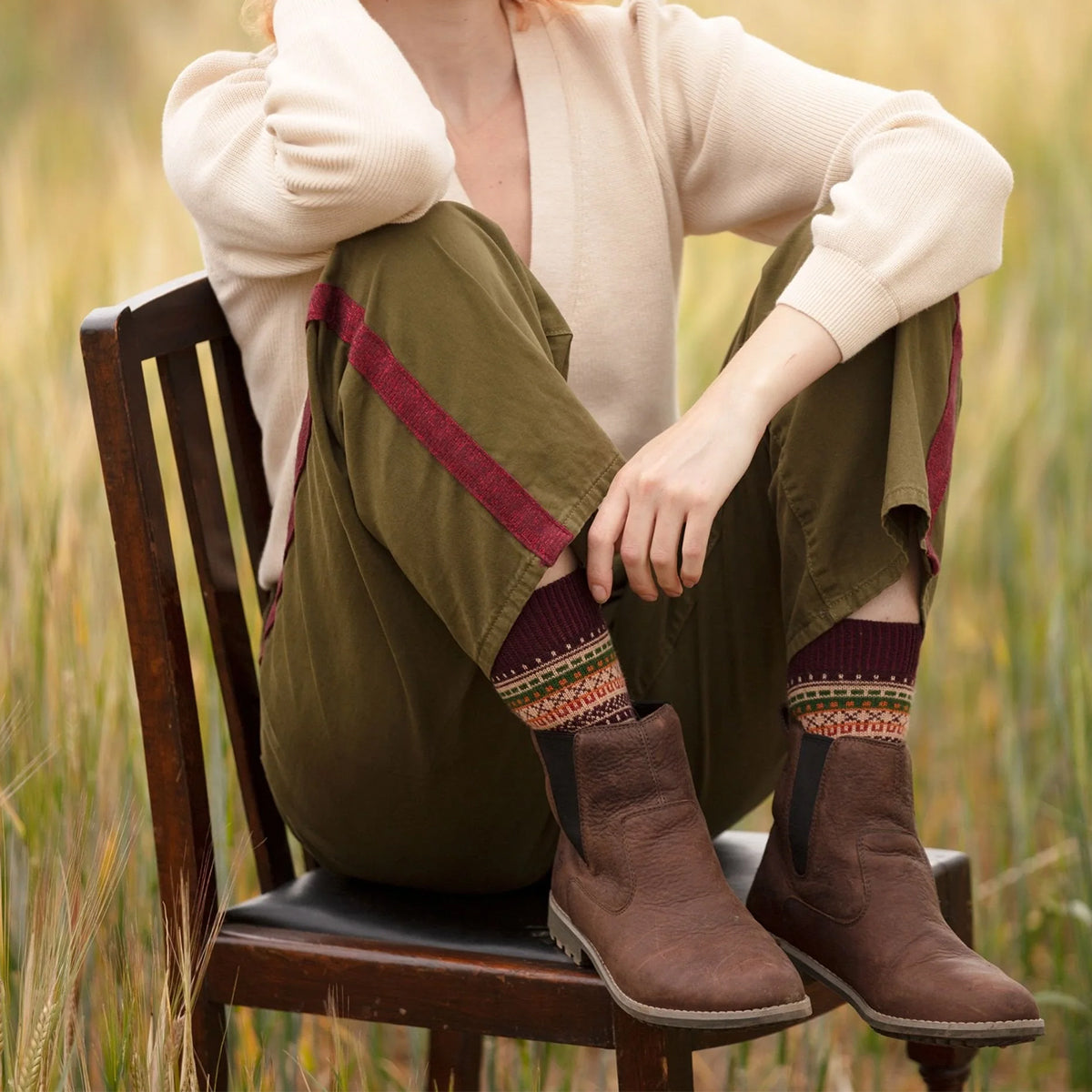 A person sitting on a wooden chair outdoors in a grassy field, wearing olive green pants with burgundy side stripes, a cream cardigan, burgandy multicolour fairisle design wool socks.