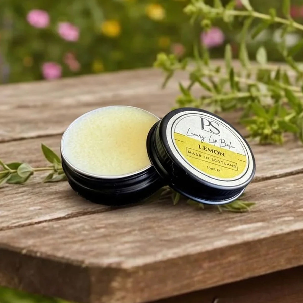 An open round tin of lemon lip balm sits on a rustic wooden surface with the lid placed beside it. The balm inside is pale yellow, and the label reads 'PS Living Lip Balm' and 'LEMON'.