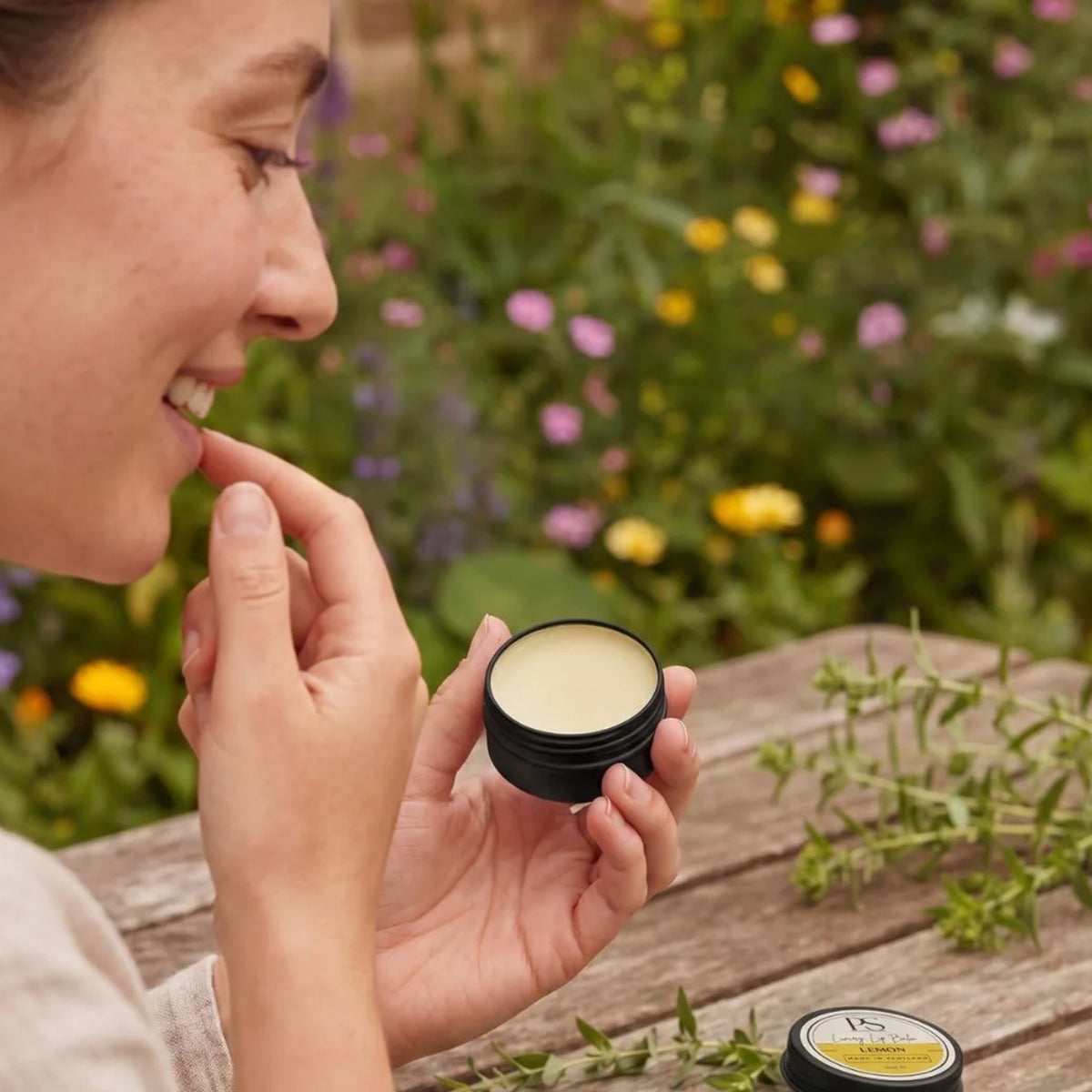 A woman outdoors on a wooden table holds an open black tin of lip balm and applies a small amount to her lips, with a blurred garden background.