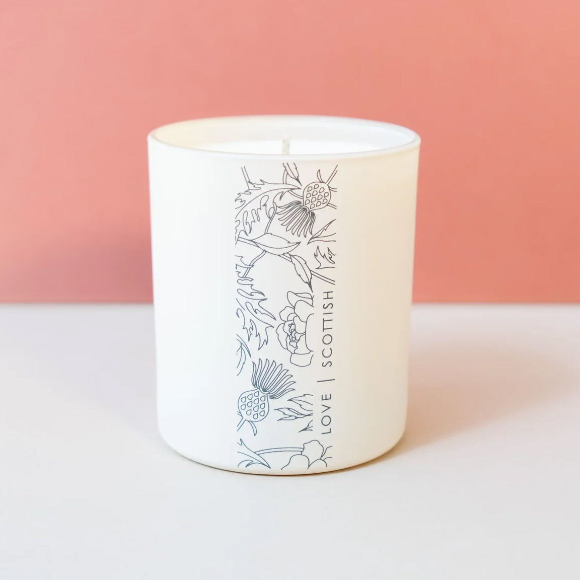 A white large glass jar candle with a single wick, featuring a vertical black line art illustration of Scottish thistles and the text 'LOVE SCOTTISH' printed on the front. The candle is set against a split background of a warm pink upper wall and a light gray lower surface.