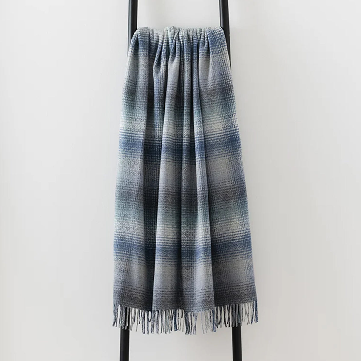 A fringed throw blanket with a blue, gray, and white striped pattern, draped over a black metal stand against a plain white wall.