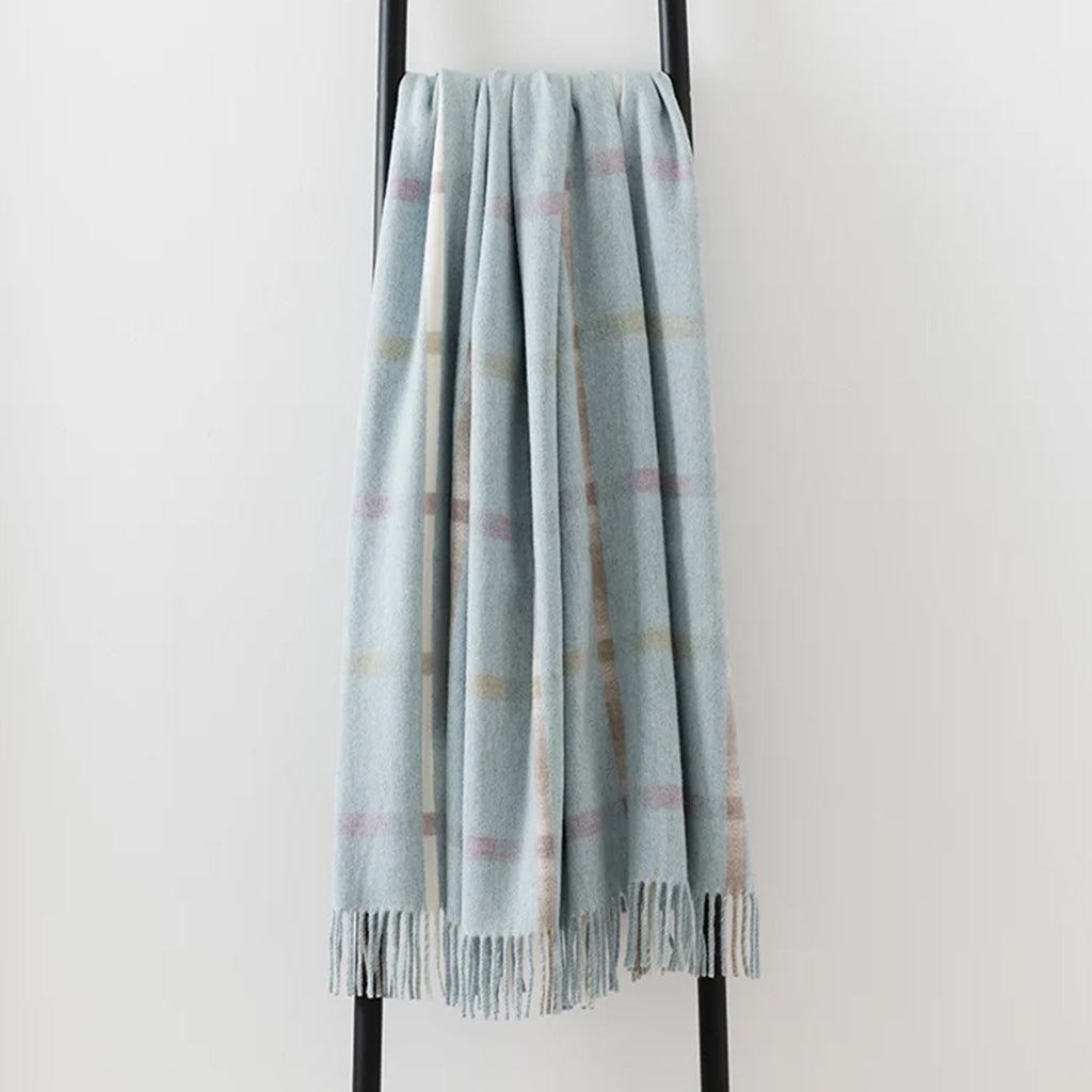 A 100% merino lambswool Remony throw draped over a black metal stand - in a pale blue shade with an elegant fringe edge.