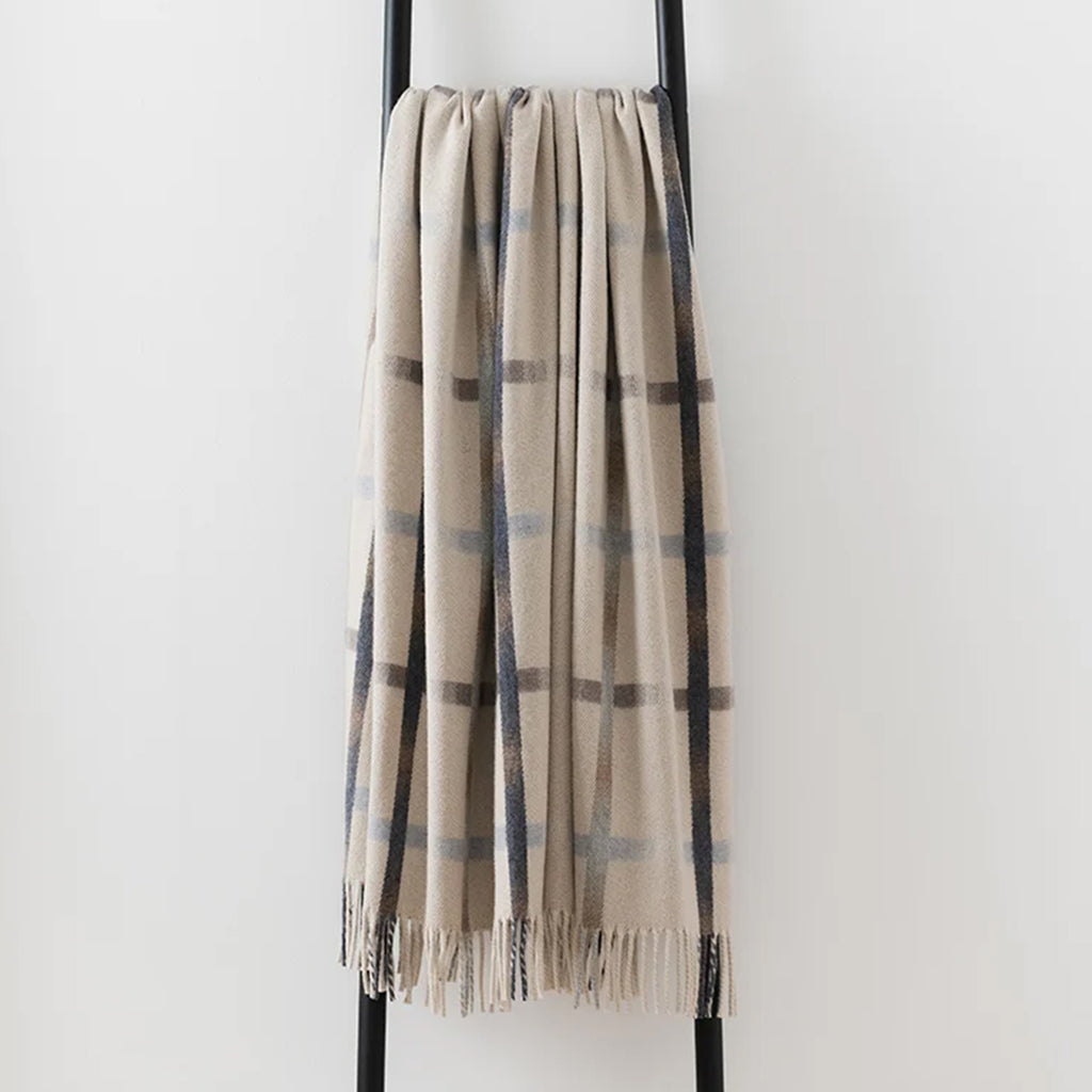 A merino lambswool woven Remony throw in shades of cream, beige and brown, draped over black metal stand. The blanket features a fringe edge.