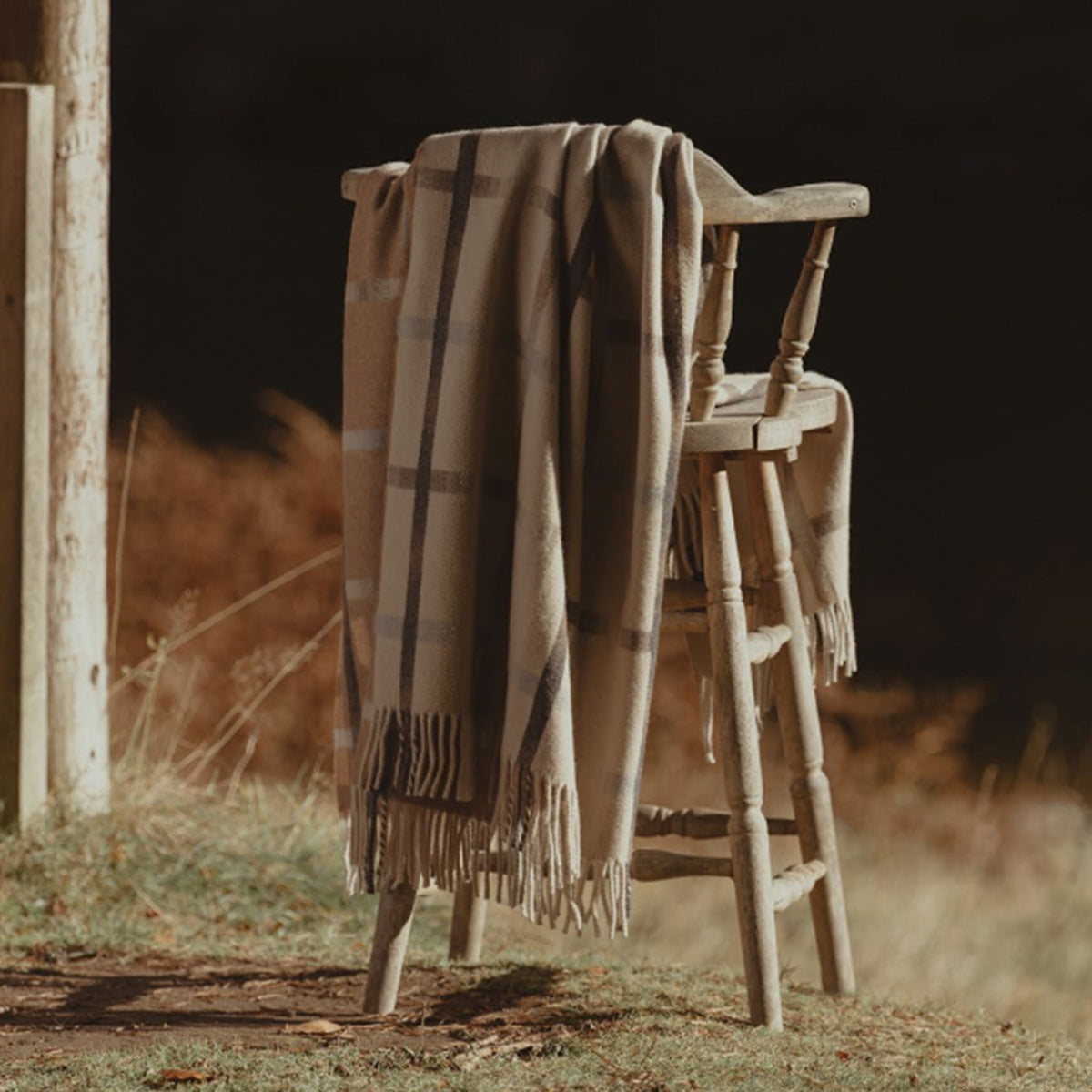 A beige plaid throw blanket with fringe edge is draped over a rustic wooden chair in an outdoor setting with warm, dry grass and a dark background.